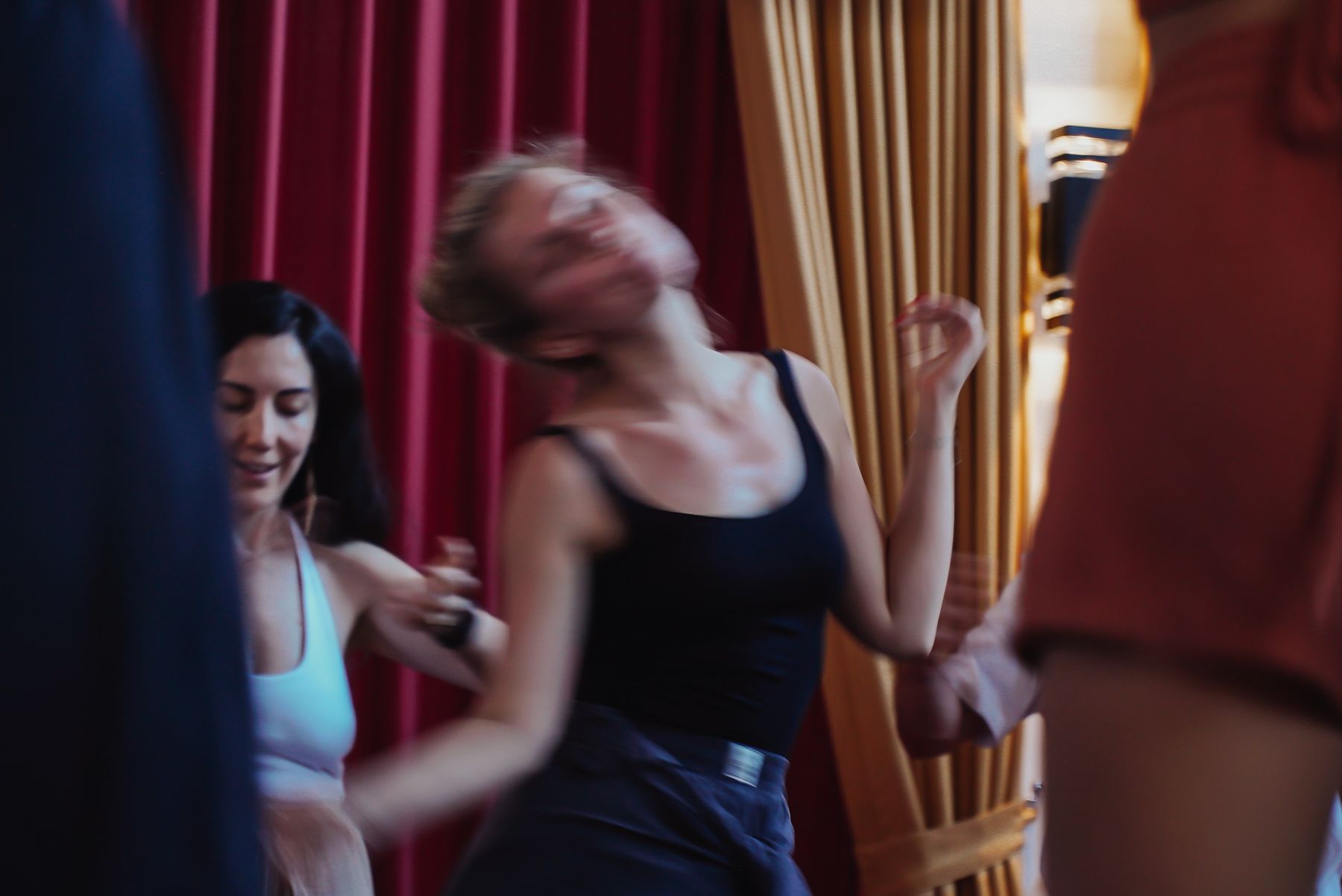 Two women dancing, eyes closed, in front of red and gold curtains.